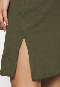 Sukienka w oliwkowym odcieniu, wykonana z miękkiego, teksturowanego materiału, charakteryzuje się bocznym rozcięciem i prostym wykończeniem, prezentując subtelne przeszycia wzdłuż krawędzi.