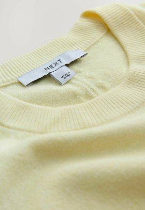 Lichtgeel gebreide trui met een geribbelde ronde hals. Label vermeldt "NEXT" als merk en "Made in China." Zachte textuur, geen zichtbare patronen.