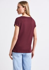 Camiseta de manga corta burdeos con puños remangados, tejido suave, corte relajado y escote redondeado, combinada con un vaquero azul claro.