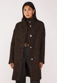 Cappotto marrone strutturato con colletto alto e chiusura a bottoni. Bottoni grandi e notevoli e una vestibilità rilassata. Indossato sopra un dolcevita scuro e pantaloni.