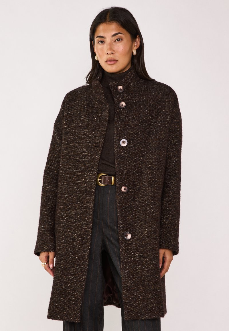 Cappotto marrone strutturato con colletto alto e chiusura a bottoni. Bottoni grandi e notevoli e una vestibilità rilassata. Indossato sopra un dolcevita scuro e pantaloni.