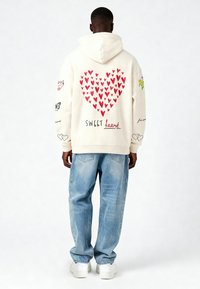 Sudadera crema con un gran gráfico de un corazón rojo en la espalda, con el texto bordado 'SWEET heart' y varios gráficos pequeños en las mangas. Llevada con jeans azules.