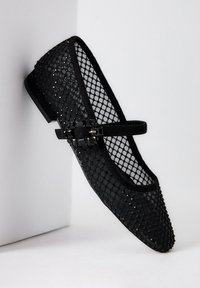 Bailarina de malla negra con puntera estructurada, detalle de hebilla decorativa y suela plana. El material tiene una apariencia texturizada y con patrón.