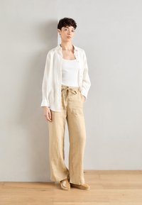 Lightweight beige linen pants with a drawstring waist, paired with a white ribbed tank top and an open white linen shirt.