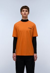 Krátký rukáv, oranžové tričko, bavlněný materiál, kulatý výstřih, s černým logem na hrudi. Nosené přes černou halenku s dlouhým rukávem.