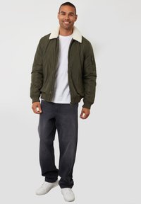 Olivgrüne Bomberjacke mit einem cremefarbenen Kunstfellkragen, Reißverschluss vorne und gerippten Bündchen, kombiniert mit grauen Jeans und weißen Sneakern.