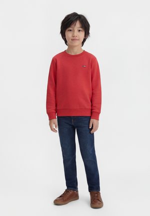 Chłopiec o czarnych włosach, ubrany w czerwoną bluzę, ciemnoniebieskie dżinsy i brązowe trampki, stojący na jasnym, jednolitym tle.