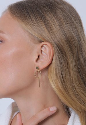 Femme blonde à la peau lisse portant une boucle d'oreille géométrique dorée avec un cercle et une barre verticale, touchant son cou avec une main manucurée.