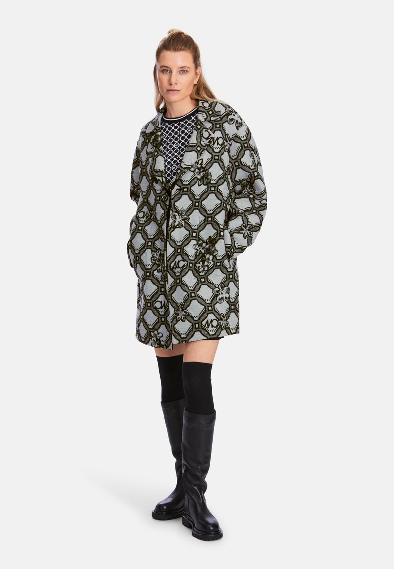 Langer gemusterter Mantel in Grau und Schwarz mit einem abstrakten geometrischen Design. Das Model trägt knielange schwarze Stiefel und ein gestricktes Oberteil darunter.