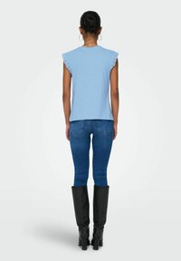 Helles blaues Kurzarmoberteil mit Spitzenbesatz, kombiniert mit blauen Denim-Jeans und schwarzen knielangen Stiefeln. Die Rückansicht zeigt eine taillierte Silhouette.