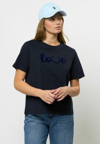 Navy bombažna majica s kratkimi rokavi ima temno modro grafiko "ljubezen" in cvetlične detajle. Kombinirana je z svetlo modro kapo in jeans hlačami.