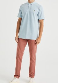 Polo shirt bleu clair avec un col, trois boutons et un petit logo noir. Assorti à un pantalon rose ajusté et des baskets blanches.