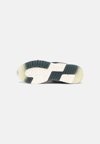 Sole di sneaker con battistrada in gomma bianca e blu scuro, caratterizzata da croci e modelli di presa texturizzati, fotografata su uno sfondo grigio chiaro.