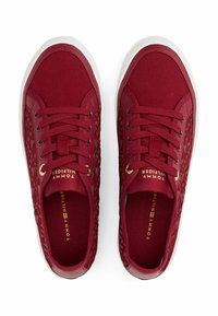 Zapatillas de lona rojas con textura tejida en los laterales, que cuentan con acentos dorados en el logo y suelas de goma blanca. Cuerdas redondas para un ajuste seguro.
