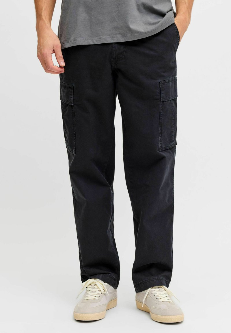Mann trägt schwarze Cargohosen mit seitlichen Taschen, graues T-Shirt und beige Sneaker, steht mit einer Hand in der Tasche.