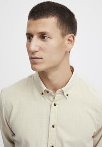 Chemise beige à boutons en tissu texturé, avec un col et des boutons foncés, portée par une personne aux cheveux courts et portant une petite boucle d'oreille.