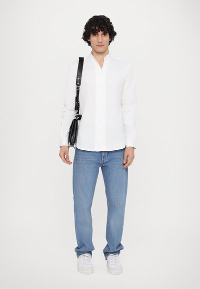 Hvit skjorte med lange ermer og knapper, lyseblå jeans og hvite joggesko. Modellen holder en svart crossover-veske med stropp. Enkel, minimalistisk design.