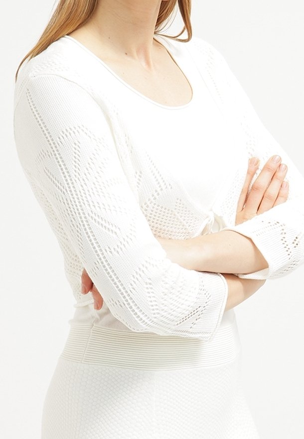 Vit kortärmad cardigan med långa ärmar, som har ett stickat mönster och rund halsringning. Materialet verkar lätt och texturerat.