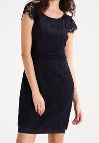 Robe en dentelle bleu marine avec une silhouette ajustée, des manches scallopées et une doublure lisse. Motifs floraux complexes sur l'ensemble du design.