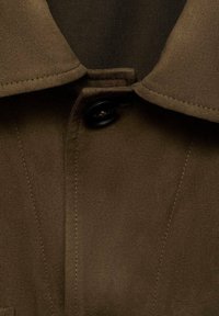 Gros plan sur le col d'un manteau marron, avec un bouton noir, une texture lisse et des coutures propres le long du bord, mettant en valeur un design structuré.