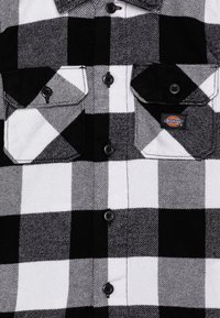Črno-bela karirana flanelna srajca z dvema sprednjima žepoma, z gumbom za zapiranje in etiketo Dickies na levem žepu.