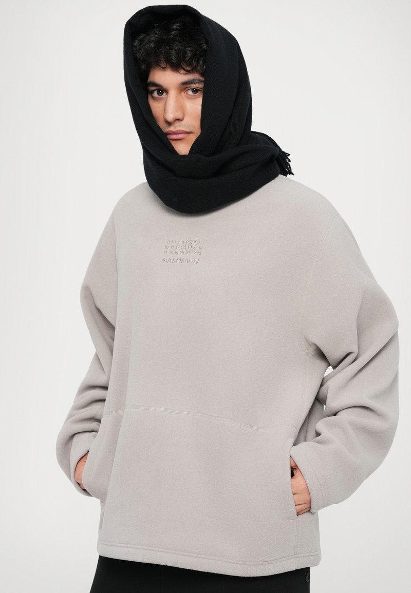 Light gray fleece sweatshirt with a relaxed fit, featuring a front pocket and small logo detail. Black scarf wrapped around the head.