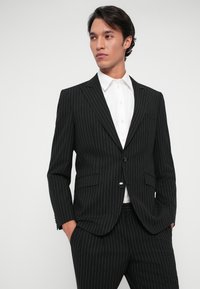 Μαύρο κοστούμι με ρίγες και κομμένη εφαρμογή. Η ζακέτα έχει κολάρο με εγκοπή και ένα κουμπί. Λευκή πουκάμισο από κάτω, με λεία υφή.