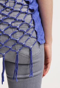 Top de tela azul con diseño de malla abierta en la parte posterior, con acentos anudados. Lleva un estilo casual combinado con unos jeans de mezclilla gris.