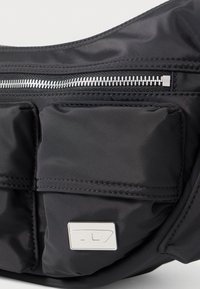 Fekete nejlon táska két első zsebbel, ezüst cipzárral és fém logó táblával. Simább textúra, lekerekített forma, minimalista dizájn.