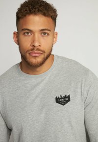 Graues Langarmshirt aus Baumwolle mit Rundhalsausschnitt, featuring ein schwarzes grafisches Logo auf der Brust, das Bäume darstellt.
