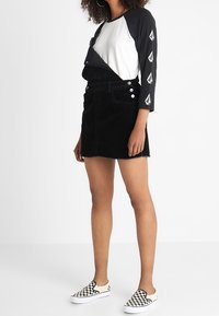 Svart kordflorskjol med knappar framtill, i kombination med en långärmad vit och svart skjorta med triangulära detaljer på ärmarna. Svart och vit rutmönstrad slip-on sko.