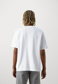 Person med kort, blond fletning iført en ensfarvet hvid t-shirt og grå bukser, stående med ryggen til mod en lys baggrund.