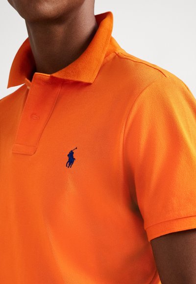 Polo orange en tissu texturé, avec un logo brodé bleu sur le côté gauche de la poitrine et un col classique.