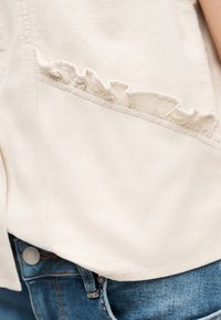 Ελαφρώς μπεζ τοπ με διαγώνια λεπτομέρεια βολάν, φορεμένο πάνω από μπλε τζιν, με εμφανή κουμπιά μπροστά και ραφές.