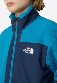 Μπλε μπουφάν από φλις με ναυτικό εξωτερικό ύφασμα, ψηλό γιακά, πλήρης φερμουάρ και λευκό λογότυπο στο στήθος. Διαθέτει υφή φλις και ομαλή ανάμειξη υφάσματος.