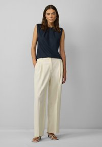 Marineblauw mouwloos top met plooien, gecombineerd met crèmekleurige high-waisted broeken en witte strappy hakken, op een grijze achtergrond.