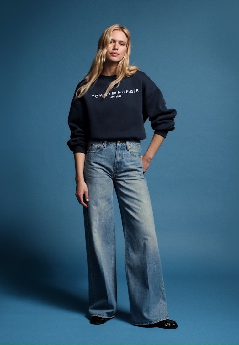 Русокоса жена стои пред син фон, носейки черна суитшърт на Tommy Hilfiger и светлосини дънки с широк крачол и черни обувки.