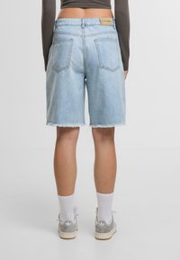 Persona vestida con pantalones cortos de mezclilla de color azul claro con dobladillos deshilachados, una camiseta de manga larga gris de corte crop, calcetines blancos y zapatillas grises, de pie contra un fondo blanco.