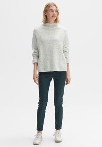 Hellgrüner Strickpullover mit hohem Kragen, kombiniert mit dunkelblauen Hosen und weißen Sneakers. Der Pullover hat ein strukturiertes Design.