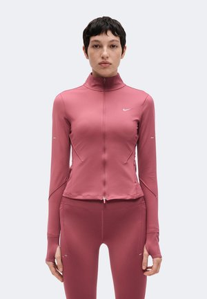 Person mit kurzen dunklen Haaren, die eine eng anliegende rosarote Reißverschlussjacke und passende Leggings vor einem einfarbigen hellen Hintergrund trägt.