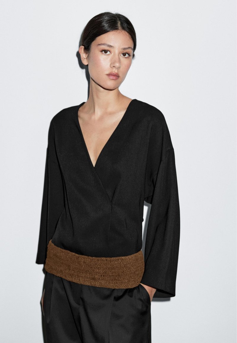 Black wrap top with a deep V-neck and oversized sleeves, featuring a textured brown waistband. Smooth fabric with a relaxed fit.