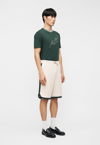 Άνδρας που φορά πράσινο κοντομάνικο πουκάμισο με λευκό κείμενο, μπεζ σορτς με πράσινη επένδυση, λευκές κάλτσες και μαύρα μοκασίνια, στέκεται μπροστά σε λευκό φόντο.