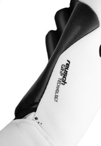 Biała rękawica z czarnym akcentem i teksturowaną powierzchnią chwytu, z etykietą "reusch GRIP TECHNOLOGY", głównie zaprojektowana do użytku sportowego.