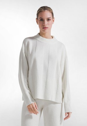 Ung kvinde med bagstrøget hår iført en hvid strikket sweater og matchende hvide bukser foran en ensfarvet hvid baggrund.