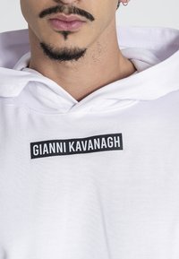 Mann trägt einen weißen Hoodie mit dem Text "GIANNI KAVANAGH" in einem schwarzen Rechteck auf der Brust, sichtbares unteres Gesicht mit gestutztem Bart und Schnurrbart.