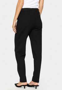 Black stretch pants with a smooth texture, elastic waistband, tapered legs, and side seams, paired with black heeled mules.
