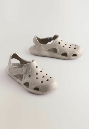 Světle šedé slip-on boty s ventilačními otvory, hladkého gumového materiálu a páskem pro bezpečné uchycení. Okrrouhlá špička a konturová podešev.