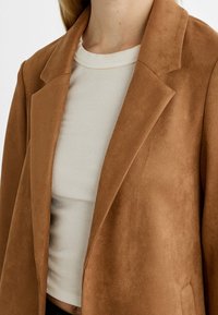 Persona vestida con una camiseta de canalé color beige claro debajo de una chaqueta de ante marrón, mostrando el torso superior y el cuello contra un fondo blanco.