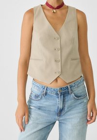 Beżowy, prążkowany top na ramiączkach z dekoltem w kształcie litery V, pięcioma guzikami i bocznymi kieszeniami, w połączeniu z jasnoniebieskimi dżinsami z przetarciami. Widoczny dodatek w postaci naszyjnika choker.