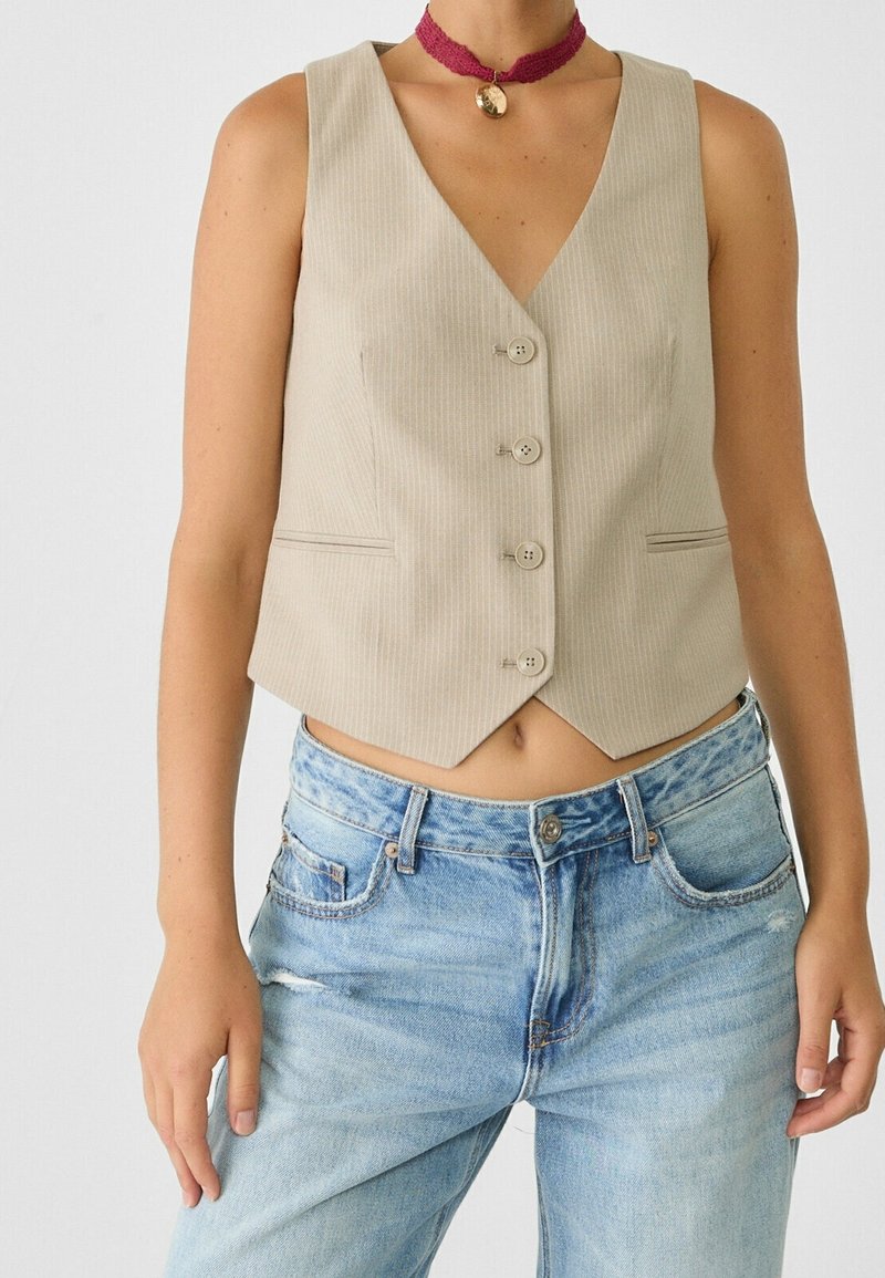 Beżowy, prążkowany top na ramiączkach z dekoltem w kształcie litery V, pięcioma guzikami i bocznymi kieszeniami, w połączeniu z jasnoniebieskimi dżinsami z przetarciami. Widoczny dodatek w postaci naszyjnika choker.